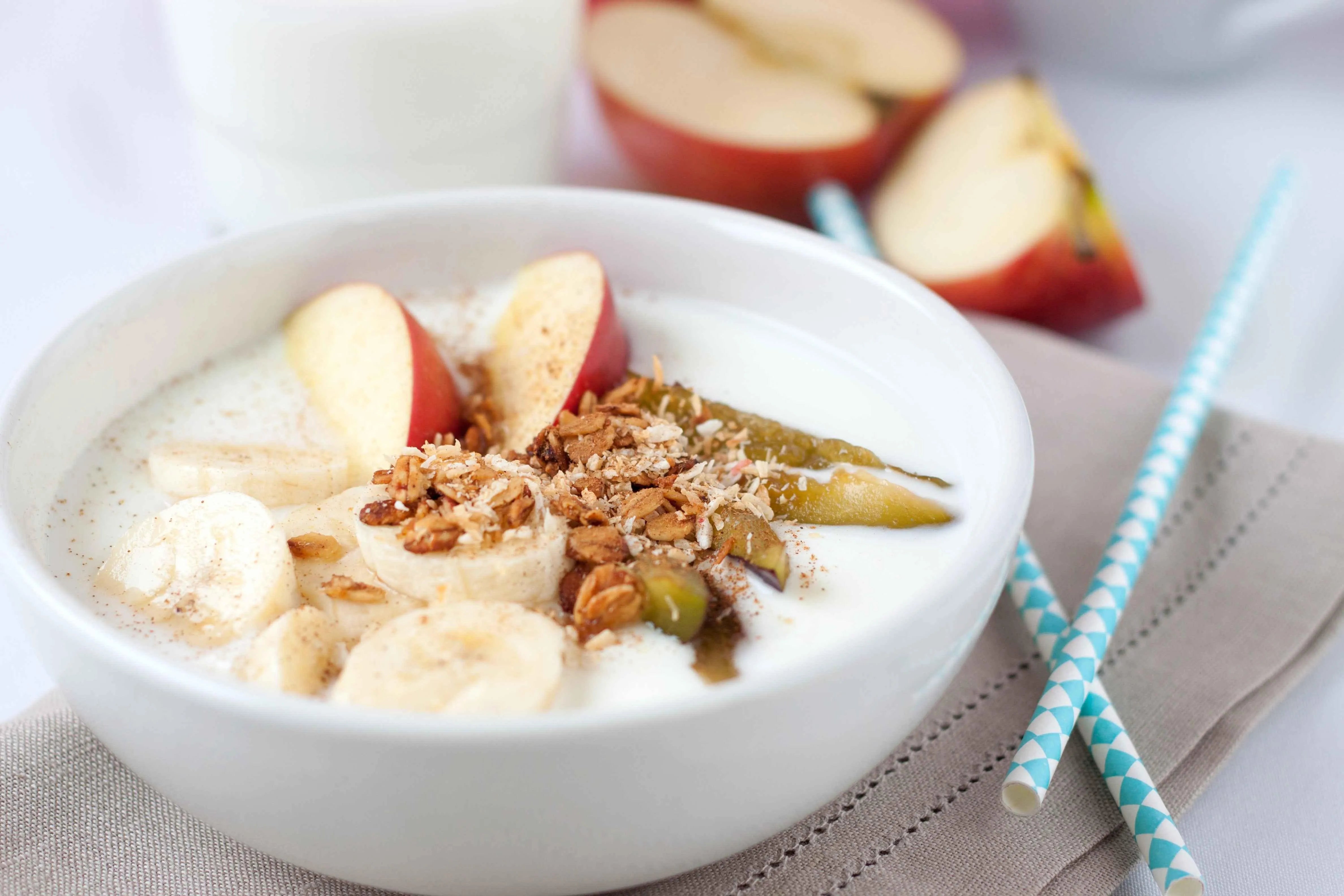 Bowl of yogurt topped with banana slices, apple wedges, granola, and green fruit